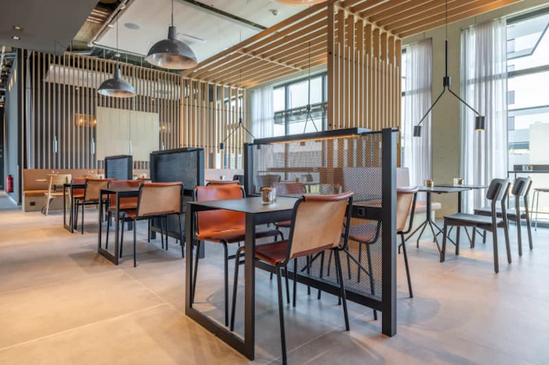 Moderne Hotelgastronomie mit Tischen, Lederstühlen, Trennwänden aus Metall und Holzlamellen vor großen Fenstern.